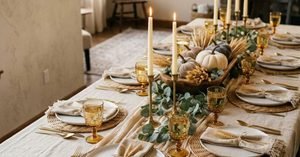 Neutral Thanksgiving table decor with a linen tablecloth, cheesecloth runner, rattan placemats, amber glasses, brass candlesticks with ivory tapers and a wooden dough bowl centerpiece with velvet pumpkins and dried wheat.